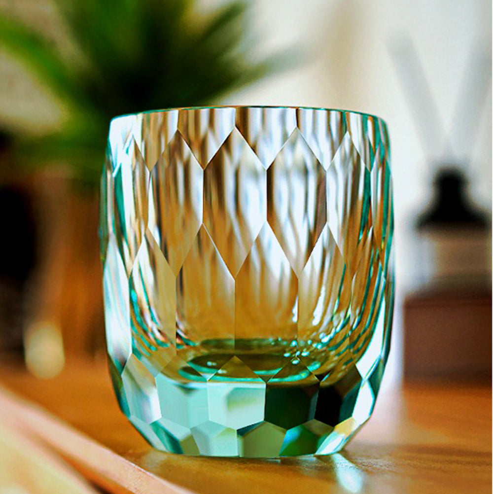 An Edo Kiriko inspired diamond-cut crystal glass with soft teal ambient reflections, sitting on a wooden counter with blurred green plant background.