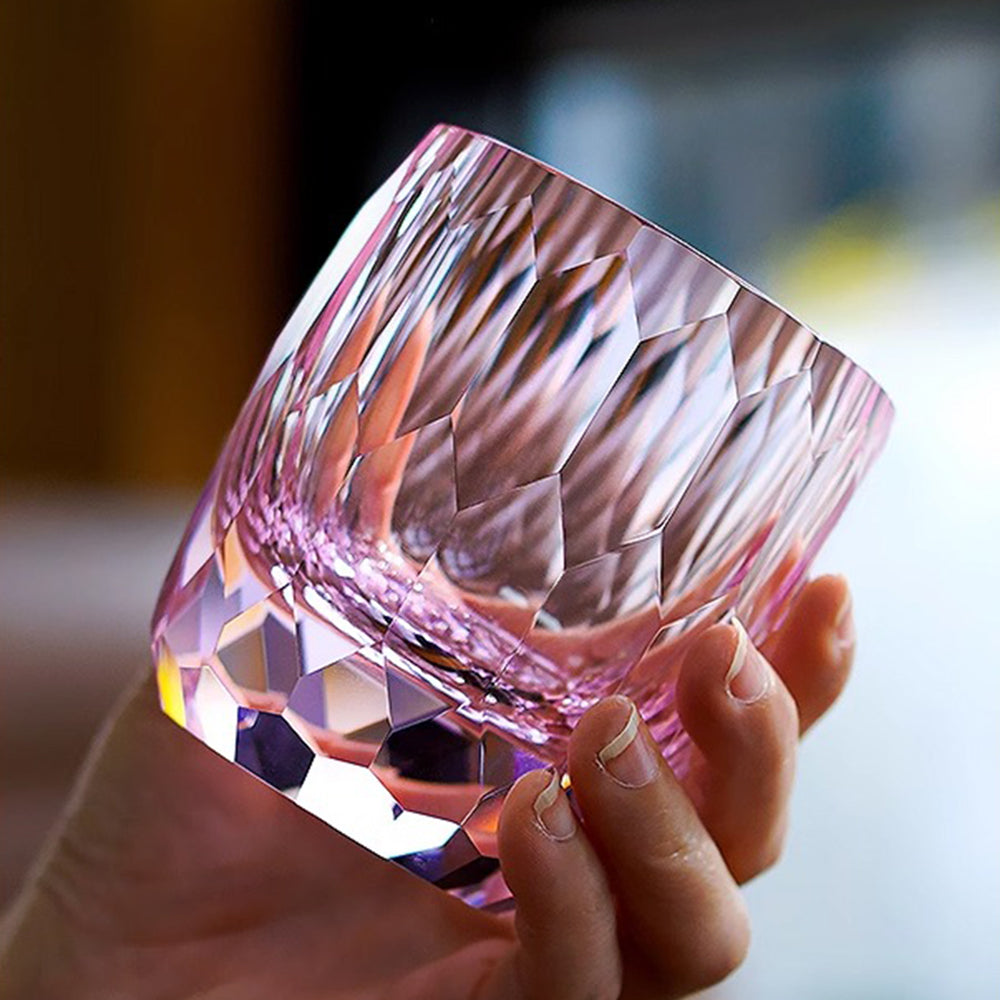 A hand holding a thick-bottomed glass tumbler with intricate geometric diamond cuts glowing in pink and purple tones.
