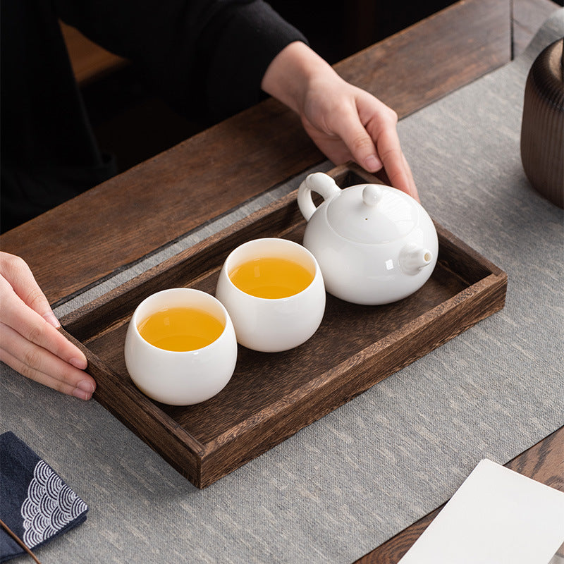 Top-down close-up of two hands serving dark Pu-erh tea from a Dehua Mutton Fat Jade Xishi teapot into matching cups on a natural wooden tea tray.